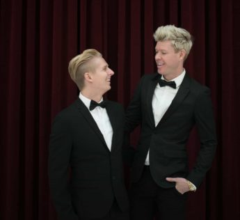 Two men in tuxedos standing in front of a red curtain.