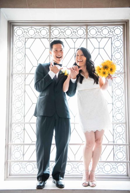 A bride and groom pose for a picture in front of a window.